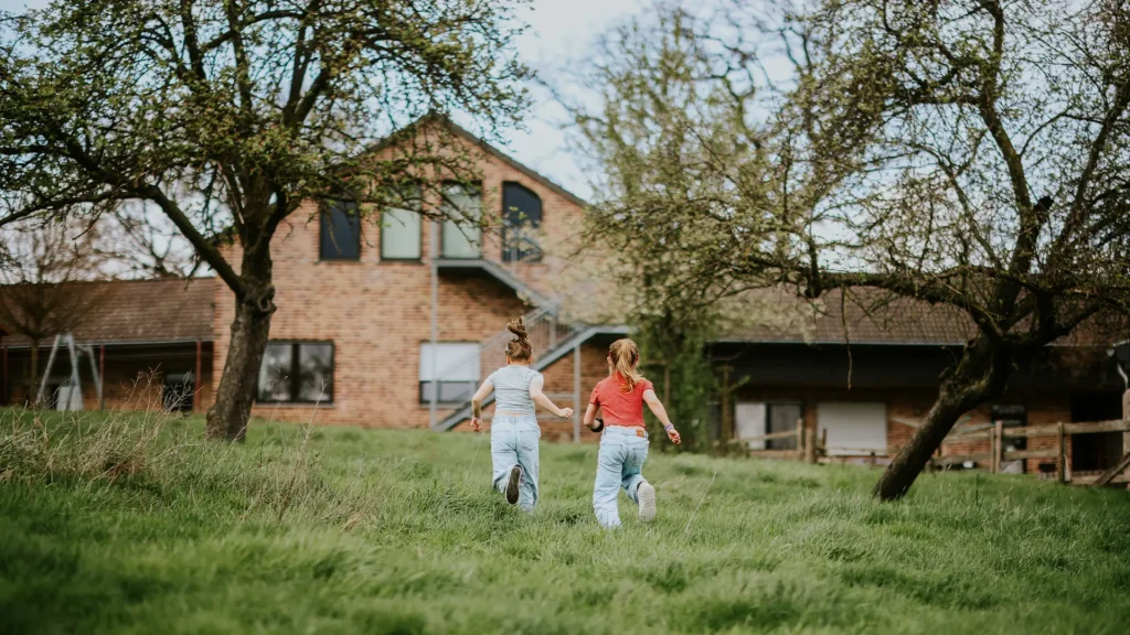 Zwei Kinder rennen über eine Wiese zu einem Haus