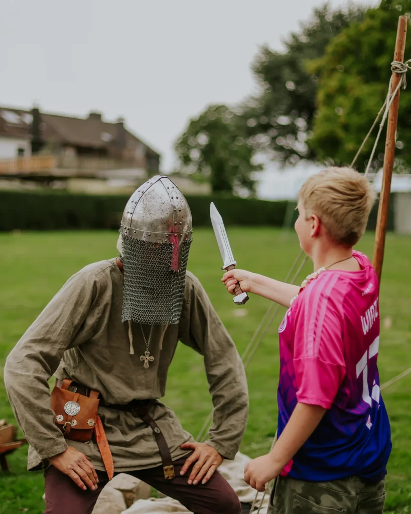 Kind hält einen Dolch und richtet ihn auf einen Erwachsenen Mann mit Ritterhelm